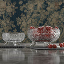 Two crystal bowls with red fruit on a reflective surface against a floral wallpaper background