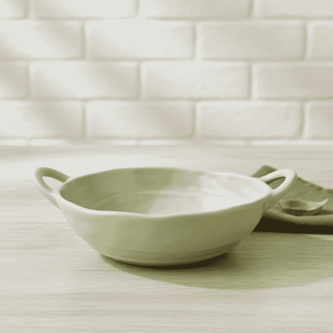 Green ceramic bowl with handles on a light wooden surface with a white tiled wall background