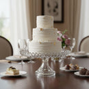 White wedding cake with gold accents on a clear glass stand in a dining setting.