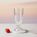 Clear glass on a white base with a strawberry next to it on a light background