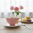 Pink teacup with saucer on a wooden table with flowers and pastries in the background