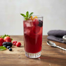 Glass of red berry cocktail with ice and mint on a wooden table with berries and a spoon.