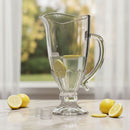 Clear glass pitcher with lemon slices on a light surface near a window with greenery outside.