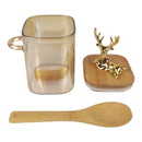 Clear mug with gold handle, bamboo spoon, and decorative deer figure on a white background