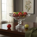 Clear glass fruit bowl with apples on a wooden table in a room with a painting on the wall.