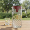 Clear glass pitcher with red lid and handle, filled with water and fruit, on a wooden table outdoors.