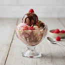 Glass sundae dish with ice cream, chocolate sauce, and raspberries on a wooden surface.