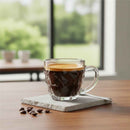 Clear glass coffee cup with a layer of foam on a wooden table, with coffee beans scattered around.