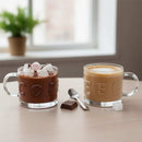 Two glass mugs, one with hot chocolate and marshmallows, the other with coffee, on a table with a window in the background.