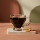 Clear glass mug filled with dark liquid on a coaster with cookies, against a warm-toned background.