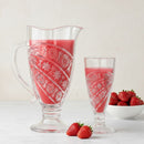 Decorative glass pitcher and glass filled with red liquid, surrounded by strawberries on a light background