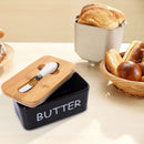Butter container with wooden lid and knife, surrounded by bread on a light surface.