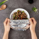 Person wrapping a dish of stir-fried vegetables and meat in tinfoil on a gray surface.