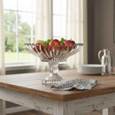 Clear glass fruit bowl with apples on a wooden table in a bright room.
