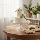 Two-tiered white wedding cake with floral decorations on a glass cake stand, accompanied by small cheesecakes on a wooden table.