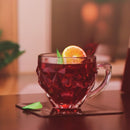 Clear glass mug filled with red liquid, garnished with an orange slice and a green leaf, on a wooden surface.