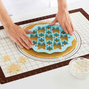 Person using a blue cookie cutter on rolled-out dough with cut-out shapes on a white surface.