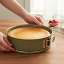 Person holding a green springform pan with a baked cake on a wooden table.